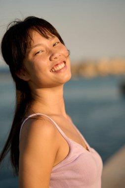 Neşeli genç bir kadının portresi deniz kenarında günbatımının tadını çıkarıyor, mutluluk ve sükunet saçıyor.