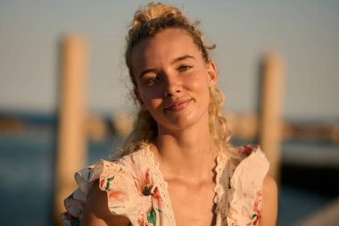 Gülümseyen sarışın kadın, sakin denizin yanında sıcak altın saatin tadını çıkarıyor, mutluluk ve rahatlığı simgeliyor.