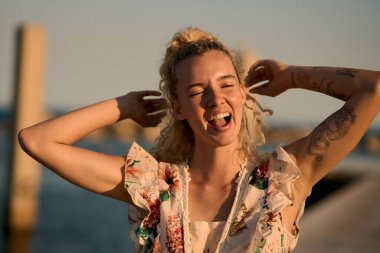 Dövmeli, kıvırcık sarı saçlı, kolları havada gülen, kumsalda altın saatin tadını çıkaran genç bir kadın.