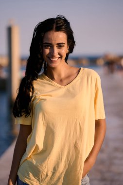 Uzun siyah saçlı, neşeli sarı tişörtlü, gün batımının sıcak altın saatinde iskelede gülümseyen genç bir kadın.