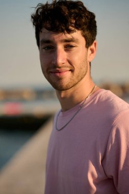 Deniz kenarında gülümseyen, pembe tişörtlü ve gümüş kolyeli şık bir gencin portresi. Günbatımının tadını çıkarıyor.