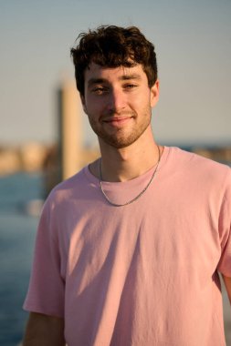 Pembe tişört giyen gülümseyen bir gencin portresi ve arka planda ortaçağ aigues mortes şehrinin gümüş kolyesi.