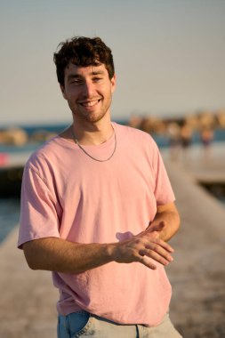 Gülümseyen genç bir adamın portresi. Elleri bir sahil kaldırımında sevinçle el kol geziyor. Gün batımının sıcacık parıltısında güneşleniyor.