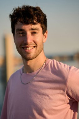 Gün batımında deniz kenarında gülümseyen pembe tişörtlü ve gümüş kolyeli neşeli bir gencin portresi.