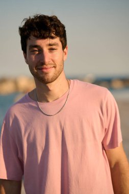 Pembe tişörtlü ve gümüş kolyeli gülümseyen genç bir adamın portresi sahilde güzel bir gün batımının keyfini çıkarıyor.