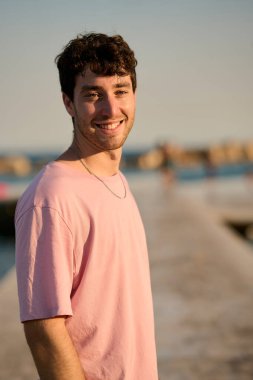 Neşeli genç bir adamın portresi plaj iskelesinde gün batımının keyfini çıkarıyor, pembe bir tişört ve gümüş bir kolye giyiyor.