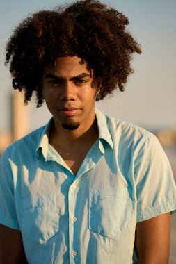 Afro saçlı, açık mavi tişörtlü, altın saatinde açık havada poz veren şık bir gencin portresi.