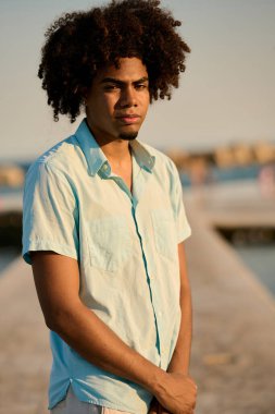 Gün batımında bir iskelede duran kıvırcık saçlı, kendine güvenen genç bir adamın portresi, altın saatin tadını çıkarıyor.