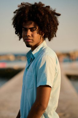 Afro saçlı genç, şık bir adamın portresi, baş döndürücü bir günbatımında bir rıhtımda güven içinde poz veriyor, rahat bir yaz havası yakalıyor.