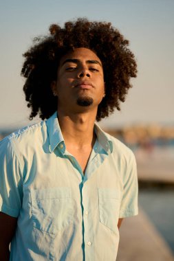 Afro saçlı şık bir gencin portresi, sakin bir yaz akşamının ılık altın saatinde güvenli bir şekilde dışarıda poz veriyor.