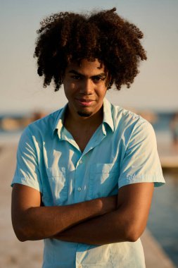 Gün batımında kumsalda kolları katlanmış gülümseyen uzun saçlı genç bir adamın portresi.