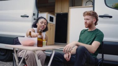 Genç, çok ırklı bir çift dinleniyor, bira içiyor, cips yiyor, karavanın dışındaki masada oturuyor.