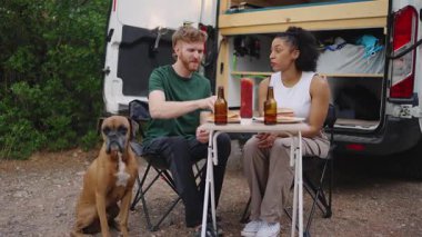 Genç, çok ırklı bir çift karavanın yanında köpekleriyle yemek yiyor ve minibüsün keyfini çıkarıyorlar.