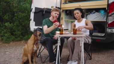 Genç bir çift ve boksör köpekleri karavanın yanındaki katlanan masada yemek yiyorlar.