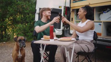 Genç çok ırklı bir çift, bira ve sosisli sandviçlerin keyfini çıkarıyor. Kamp arabalarının arkasındaki katlama masasında oturuyorlar. Yanında da boksör köpekleri var.