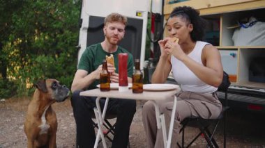 Sosisli yiyen ve yanında boksör köpeğiyle bira içen çok ırklı genç bir çift minibüslerinin önünde oturuyor.