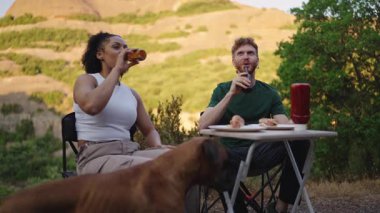 Çok ırklı genç çift kamp sırasında rahatlatıcı bir anı paylaşıyor, boksör köpekle yemek ve içeceklerin keyfini çıkarırken aynı zamanda doğa manzarasında minibüs hayatı macerası yaşıyorlar.