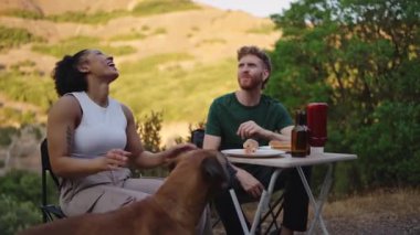 Neşeli çoklu etnik çift, altın gün batımında manzaralı macerada sadık boksör köpeği eşliğinde karavanın yanında doyurucu bir yemek paylaşıyor.