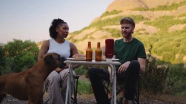 Minibüs hayatı macerası sırasında manzaralı bir dağ manzarasında boksör köpeğiyle yemek yiyen çok ırklı mutlu bir çift.