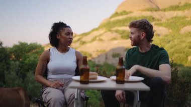 Genç çoklu etnik çiftler sohbet ediyor, bira içiyor ve dışarıda köpekleriyle sandviç yiyorlar.