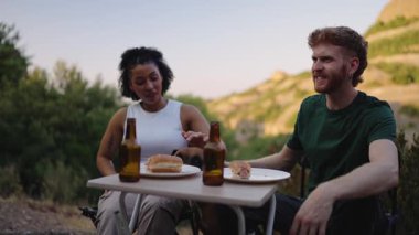 Minibüs macerası sırasında kaygısız anın tadını çıkaran, gün batımında manzaralı dağ manzarasına karşı yemek ve kahkaha paylaşan çok ırklı genç bir çift.
