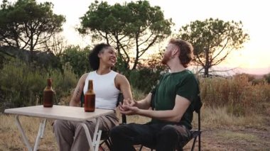 Gün batımında kamp gezisi sırasında masada oturup bira içerek gülen çok ırklı genç bir çift.