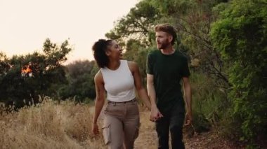 Genç çoklu etnik çiftler doğada sakin bir yürüyüş yapıyor, el ele tutuşuyor ve kahkahalarını paylaşıyorlar.