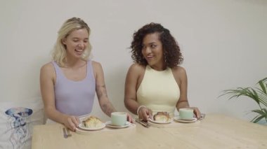 Two happy multicultural young women sitting at a table in a bright cafe, laughing and talking while having coffee and pastries together