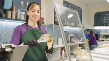 Smiling female employee at an ice cream shop scooping artisanal gelato into a waffle cone. Slow motion view of a server preparing a sweet dessert
