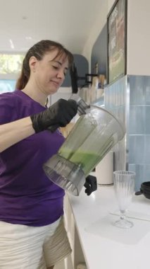 Woman in a purple shirt and black gloves carefully pouring a freshly made green vegetable smoothie from a blender into a tall glass in a kitchen