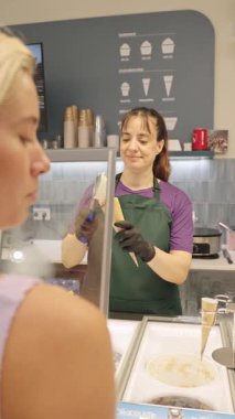Female ice cream parlor worker in an apron and gloves putting a scoop of fresh gelato onto a waffle cone for a young woman customer