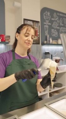 Female worker in an apron serving a scoop of vanilla ice cream into a waffle cone. Slow motion shot of a small business employee working