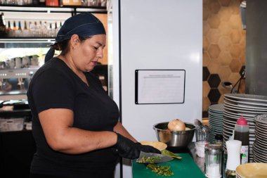 Woman chef wearing a bandanna and black gloves preparing food, chopping green vegetables on a cutting board in a professional kitchen