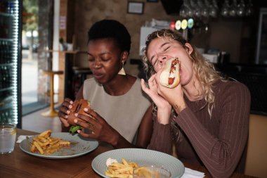 Two diverse women friends enjoying eating burgers, hot dogs, and french fries at a casual restaurant. Fast food meal