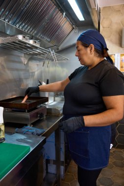 Woman cook wearing uniform and gloves preparing sausages on a griddle in a commercial kitchen, ensuring food hygiene