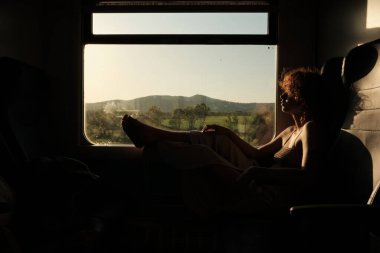 Young woman relaxing in a train at golden hour, contemplating the landscape passing by the window during her journey