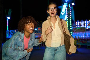 Two young women laughing, enjoying a fun night out at a vibrant amusement park, showing happiness and friendship