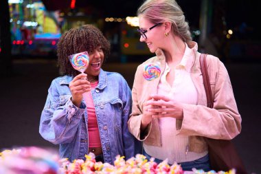 Two happy diverse friends enjoying a fun night out at an amusement park while choosing colorful lollipops from a stand