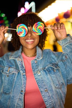 Happy woman holding rainbow lollipops over eyes, posing playfully in a denim jacket at a lively carnival fair at night