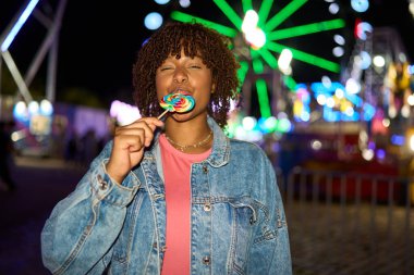 Young woman with closed eyes enjoying a sweet rainbow lollipop at night. Amusement park lights blurring in background
