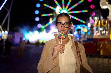 Young woman wearing glasses making a funny face while holding a rainbow lollipop at a vibrant night amusement park