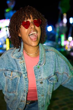 Young woman with heart sunglasses and dreadlocks expressing joy, having fun at night carnival with bright lights