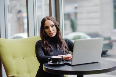 The girl is sitting at the table, drinking coffee and working on a laptop. Business woman pet coffee in the office.