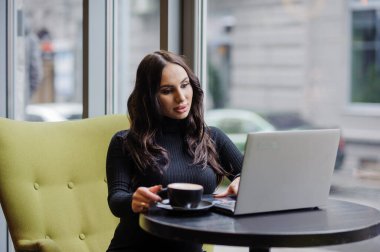 The girl is sitting at the table, drinking coffee and working on a laptop. Business woman pet coffee in the office.