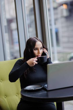 A business woman is sitting at a table in the office, talking on the phone, working on a laptop and drinking coffee.