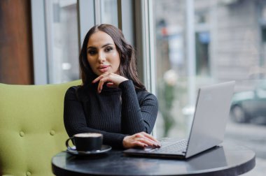 Business woman sitting at a table in the office, working on a laptop and drinking coffee.