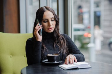 Business woman sitting at a table by the window and talking on the phone.