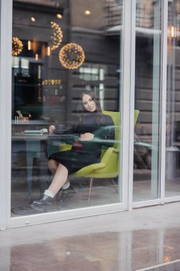 A beautiful young woman in a black dress sits in a cafe by the window and drinks coffee. Coffee break.