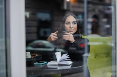 A beautiful young woman in a black dress sits in a cafe by the window and drinks coffee. Coffee break.