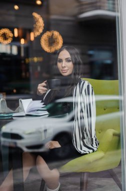 A beautiful young woman in a black dress sits in a cafe by the window and drinks coffee. Coffee break.